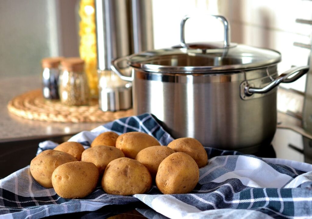 Mehrere frische Kartoffeln auf einem Küchentuch neben einem Edelstahltopf mit Kartoffelwasser auf dem Herd, bereit für die Zubereitung. Gewürzgläser und Küchenutensilien im Hintergrund.